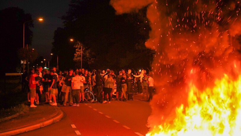 Menschen protestieren auf einer Straße, auf der ein Feuer lodert, gegen die Ankunft hunderter Geflüchteter aus Afghanistan. Die Polizei hat eine Protestaktion, zu der sich rund 250 Menschen in der Nähe des Armeelagers Harskamp (Niederlande, Provinz…