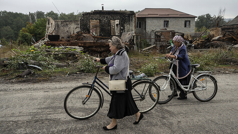 Zwei Frauen gehen mit ihren Fahrrädern auf einer Straße neben zerstörten russischen Schützenpanzer und zerstörten Häusern spazieren. Foto: Evgeniy Maloletka/AP/dpa