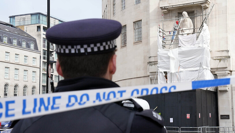 Ein Polizist sieht zu wie ein Mann, der eine Spiderman-Maske trägt, mit Hammer und Meißel eine Statue eines umstrittenen Bildhauers am Hauptquartier der BBC beschädigt. Foto: Jonathan Brady/PA Wire/dpa