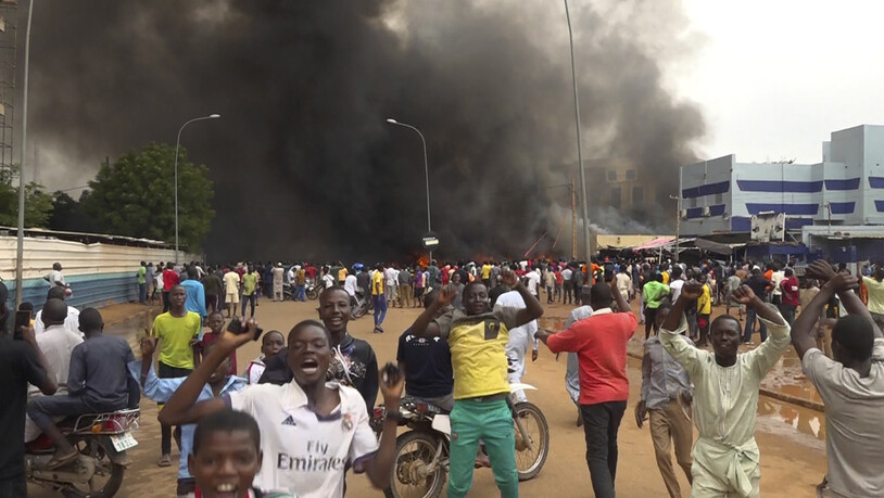 Mit dem brennenden Hauptquartier der Regierungspartei im Rücken demonstrieren Anhänger meuternder Soldaten im Niger. Foto: Fatahoulaye Hassane Midou/AP/dpa