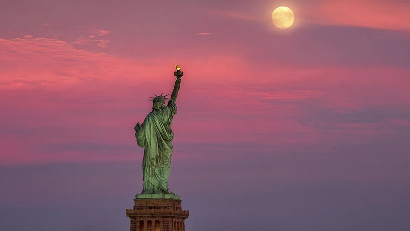 ARCHIV - Der Vollmond erhebt sich bei Sonnenuntergang hinter der Freiheitsstatue in New York. In der Millionenmetropole hat es vorläufigen Daten zufolge erstmals seit dem Ausbruch einen Tag ohne bestätigten oder wahrscheinlichen Todesfall nach einer…