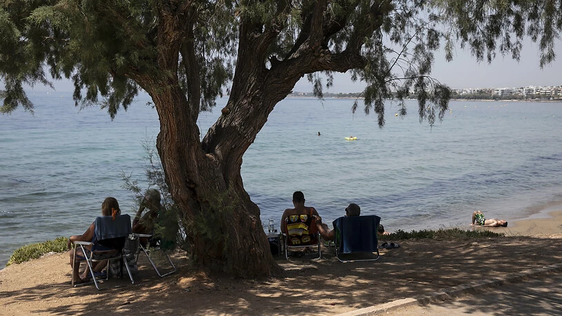 Besucher sitzen im Schatten unter einem Pinienbaum am Strand des Vororts Kavouri südwestlich von Athen. Foto: Yorgos Karahalis/AP/dpa