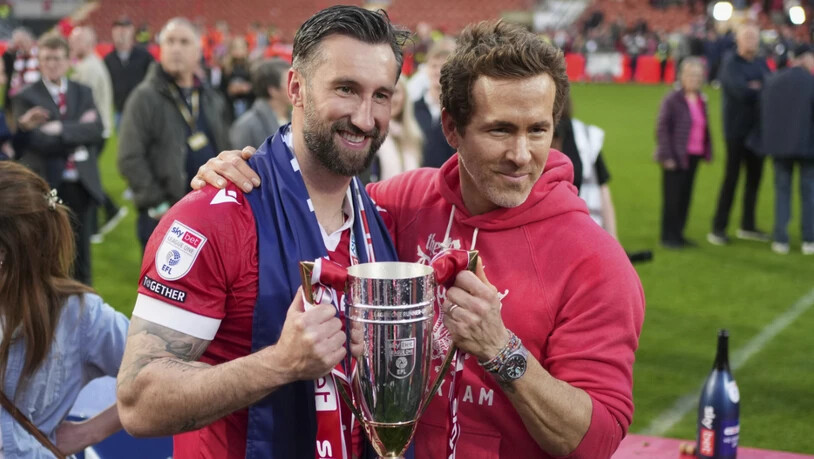 Der Hollywood-Star Ryan Reynolds, der seit gut fünf Jahren Co-Besitzer des AFC Wrexham ist, und Spieler Ollie Palmer (links) posieren nach dem Aufstieg in die Championship in der vergangenen Saison mit dem Pokal