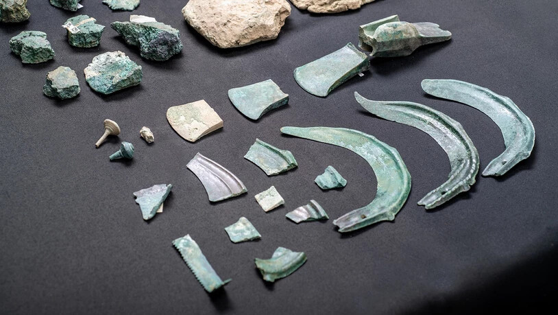 Fundstücke: Sicheln, Beile, Nadeln sowie ein Sägefragment. Im Hintergrund zu sehen sind ein Gusskuchen beziehungsweise Rohmetallfragmente aus Kupfer.